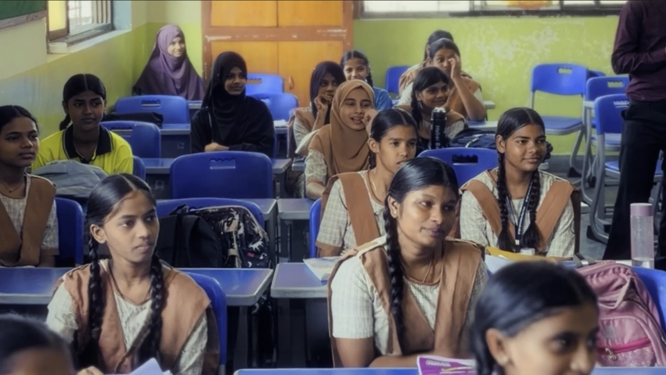 Students at desks in a government-school classroom, watching something at the front of the room