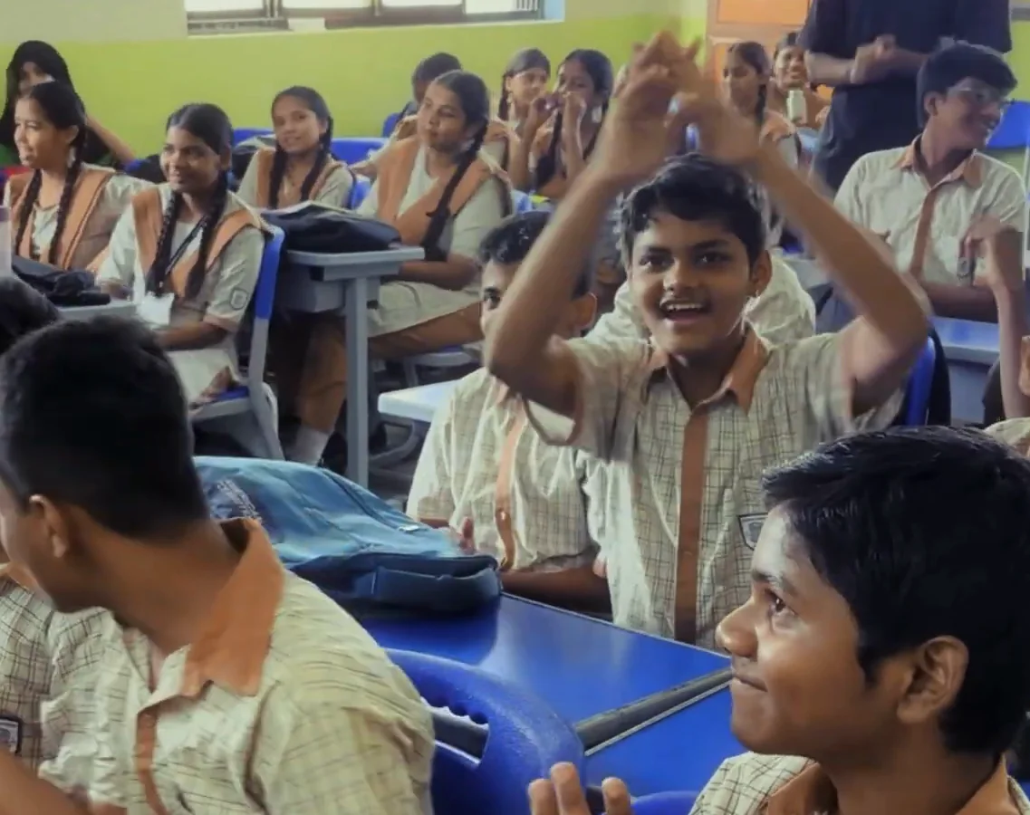 Students in their classroom mid-activity, one with both arms raised and cheering while the others laugh