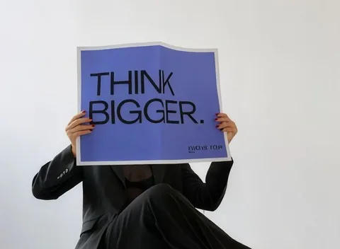 Someone seated on a stool holding a sign reading THINK BIGGER over their face