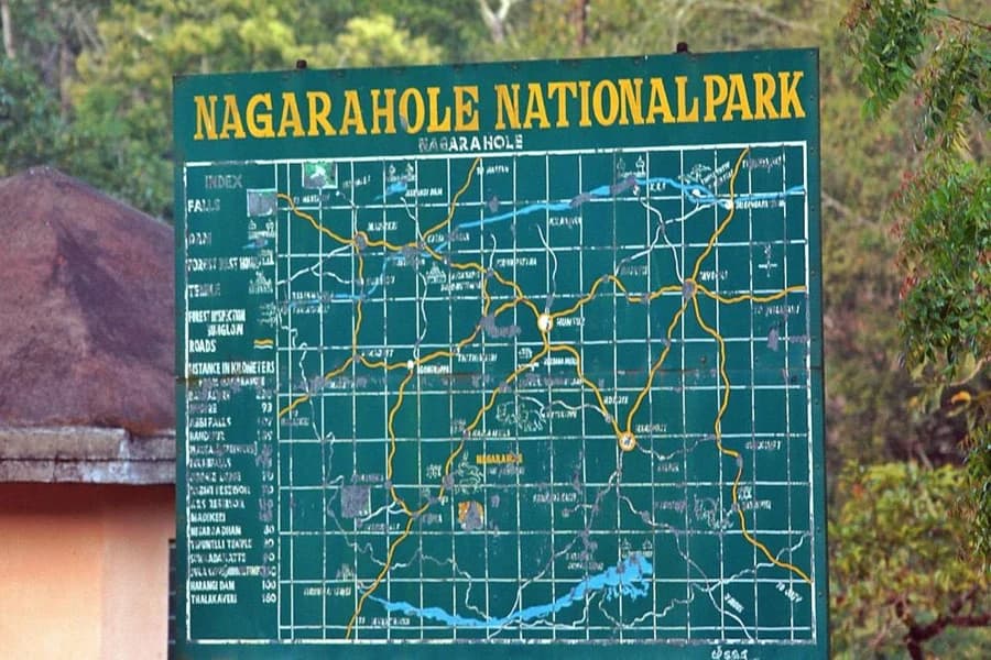 A green Nagarahole National Park signboard showing a hand-drawn grid map of the reserve's roads and an index of distances, mounted beside a forest rest-house roof
