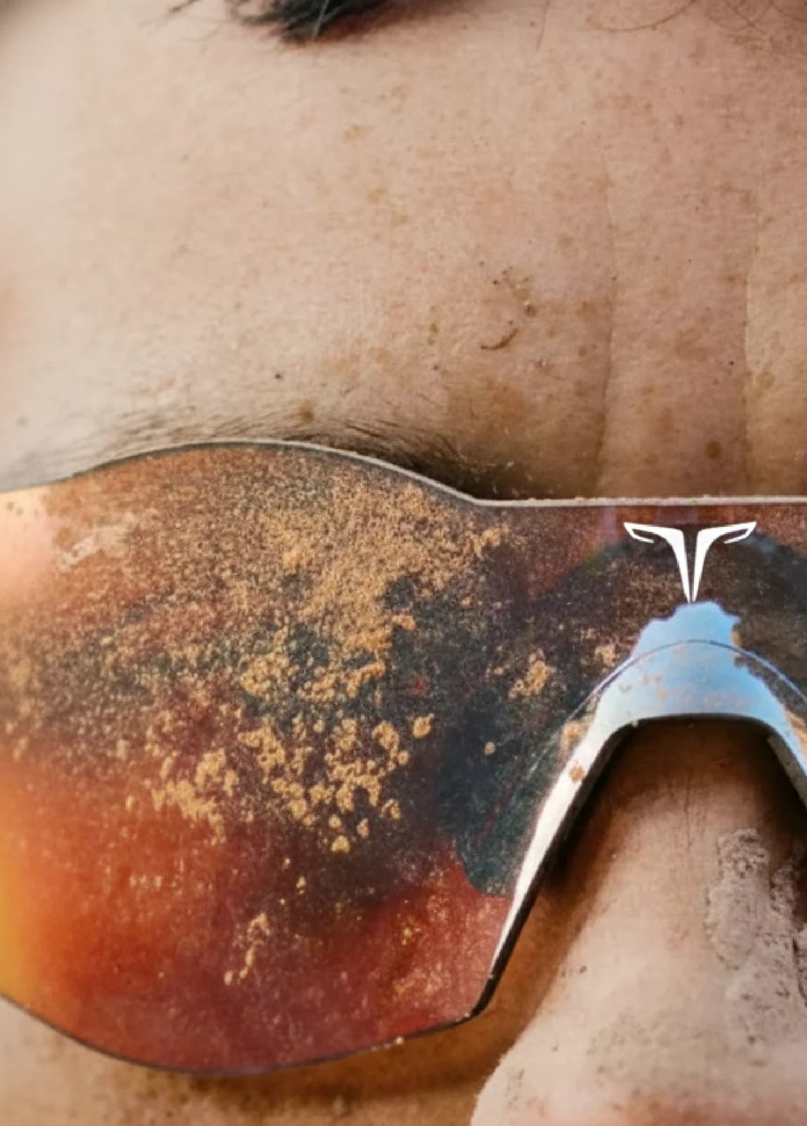 The frames worn close up, lenses caked in dust, the mark picked out in white on the brow