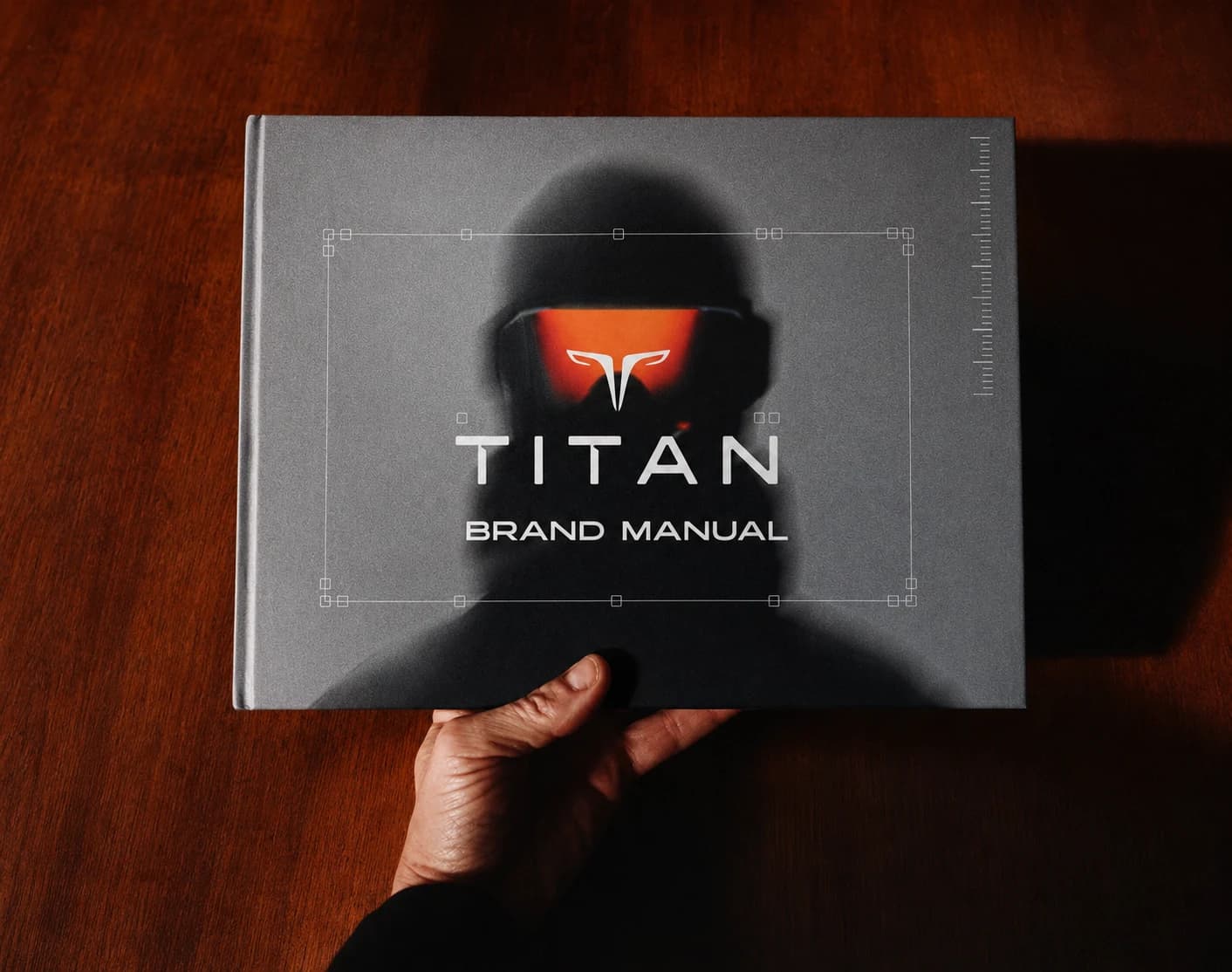 The printed TITAN brand manual held in one hand over a dark wood table